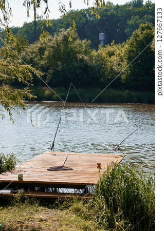 Serene Lakeside Fishing Scene: Two Rods on Wooden Dock, Green Trees, Water Reflection, Bright Daylight, Peaceful Outdoor Recreation, Summer Vibe 127667133