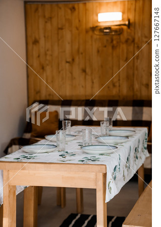 Rustic Wooden Dining Scene: Family Meal Setup with Checkered Bench, Light Wood Table, Plates and Greenery in Cozy Cabin Interior. Rustic Wooden Dining Scene: Family Meal Setup with Checkered Bench, Light Wood Table, Plates and Greenery in Cozy Cabin Interior. 127667148