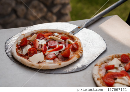 Outdoor Wood-Fired Pizza Preparation with Tomato and Mozzarella Toppings, Rustic Stone Oven, Steel Peel, Summer Scene 127667151