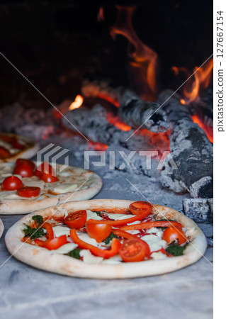 Rustic Wood-Fired Pizza with Fresh Tomatoes and Basil: Close-Up of Artisanal Pies Baking in Stone Oven with Burning Embers. Rustic Wood-Fired Pizza with Fresh Tomatoes and Basil: Close-Up of Artisanal Pies Baking in Stone Oven with Burning Embers. 127667154