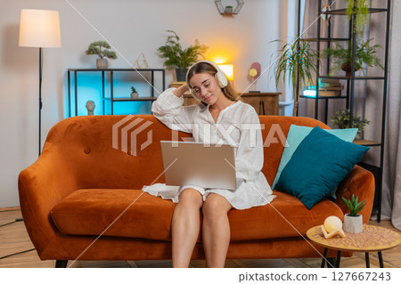 Happy overjoyed young woman girl in wireless headphones listening music sitting on sofa at home Happy overjoyed young woman girl in wireless headphones listening music sitting on sofa at home 127667243