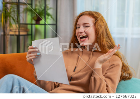 Excited young woman opening envelope letter and reading good news sitting on sofa at home apartment 127667322