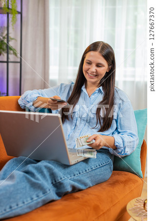 Rich happy young woman counting money dollar cash using laptop calculate domestic bills sits on sofa Rich happy young woman counting money dollar cash using laptop calculate domestic bills sits on sofa 127667370
