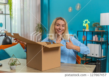 Happy blond businesswoman unpacking opening delivery parcel box purchase sits at desk in apartment 127667457