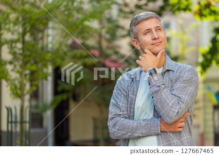 Thoughtful smiling mature man with pensive expression touching chin looking around on city street 127667465