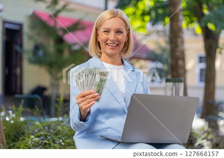 Happy mature Caucasian businesswoman CEO holding cash and using laptop computer on city street Happy mature Caucasian businesswoman CEO holding cash and using laptop computer on city street 127668157
