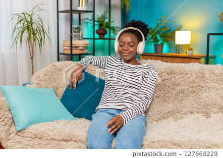 African American woman relaxing sofa eyes closed deep breaths listening music headphones indoors 127668229