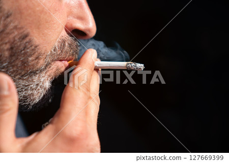Close up of bearded man holding a lighter and smoking a cigarette on black background. 127669399