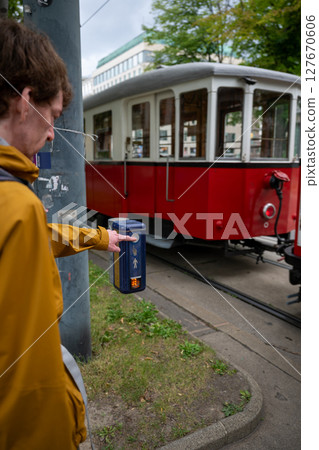 man pedestrian in yellow jacket presses pedestrian crossing button beside red tram on a city street 127670606
