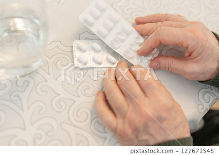 Senior person hands holding blister packs of pills over a patterned tablecloth, with a glass of water nearby, copy space. Concept of senior health care and daily medication routine Senior person hands holding blister packs of pills over a patterned tablecloth, with a glass of water nearby, copy space. Concept of senior health care and daily medication routine 127671548