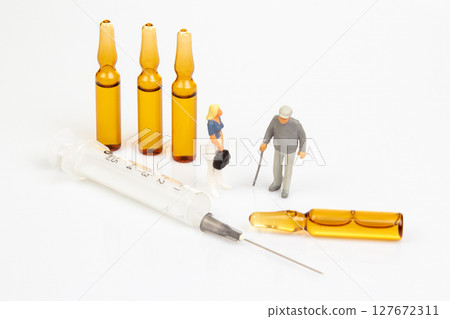 Miniature doctor and elderly figurine stand near three amber vials, a syringe, and a broken vial  127672311