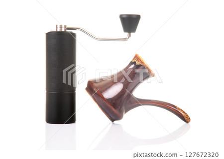 A black manual coffee grinder with a metallic handle stands next to a copper cezve A black manual coffee grinder with a metallic handle stands next to a copper cezve 127672320