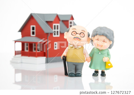 Two miniature elderly figurines, a man and a woman with a bag, stand in front of a red toy house  127672327