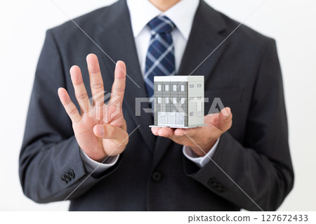 一位身著西裝的中年男性商人手持建築模型,豎起四根手指 一位身著西裝的中年男性商人手持建築模型,豎起四根手指 127672433