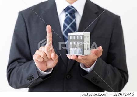 一位身著西裝的中年男性商人,手裡拿著一棟建築模型,用食指指著 一位身著西裝的中年男性商人,手裡拿著一棟建築模型,用食指指著 127672440