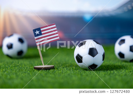 Small Liberian flag stands proudly on lush green grass field next to miniature soccer balls, symbolizing national pride and international football championship success Small Liberian flag stands proudly on lush green grass field next to miniature soccer balls, symbolizing national pride and international football championship success 127672480