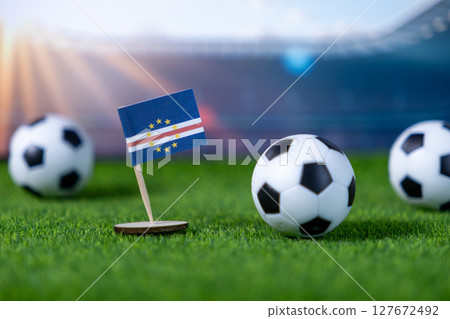A small Cape Verde flag standing proudly with soccer balls on vibrant green grass, set against a blurred stadium backdrop, symbolizing international football championships A small Cape Verde flag standing proudly with soccer balls on vibrant green grass, set against a blurred stadium backdrop, symbolizing international football championships 127672492