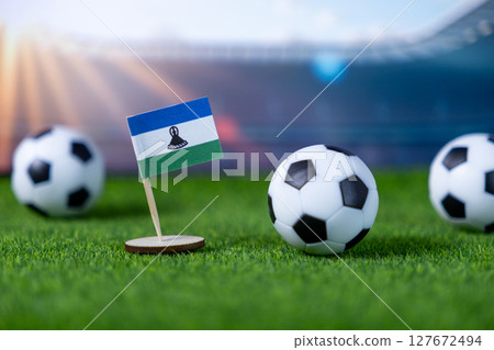 Lesotho national flag miniature prominently placed on vibrant green artificial grass with classic soccer balls, representing global football competition 127672494