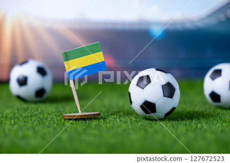 Small Gabon flag stands with mini soccer balls on green artificial grass, symbolizing global football competition and national team pride at a stadium venue Small Gabon flag stands with mini soccer balls on green artificial grass, symbolizing global football competition and national team pride at a stadium venue 127672523