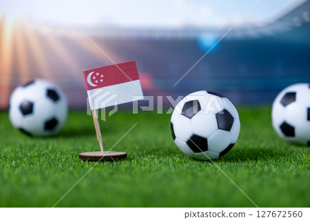Miniature Singapore national flag with several soccer balls on vibrant green grass in a blurred stadium, representing national pride and global football competition 127672560