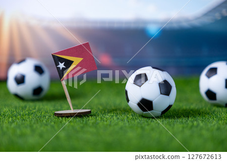Miniature Timor-Leste flag and soccer balls on green grass in a blurred stadium, symbolizing global football tournaments, national pride, and sports events Miniature Timor-Leste flag and soccer balls on green grass in a blurred stadium, symbolizing global football tournaments, national pride, and sports events 127672613