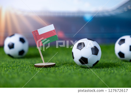 Oman national flag proudly displayed with soccer balls on vibrant green stadium grass, representing national team's participation in international football sports competition Oman national flag proudly displayed with soccer balls on vibrant green stadium grass, representing national team's participation in international football sports competition 127672617