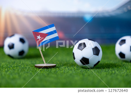A Cuban flag stands firmly on green artificial grass with miniature soccer balls, portraying national representation and excitement for international football events in a stadium setting 127672640