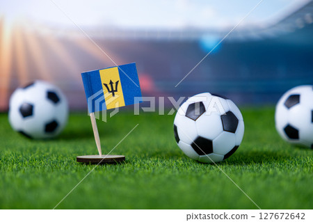 Small Barbados national flag miniature proudly stands with soccer balls on vibrant green artificial grass, symbolizing national team spirit and global football championship 127672642
