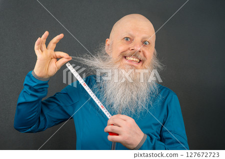 happy bald man demonstrating the length of his large white beard using a metric tape  127672723