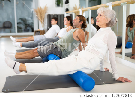 Elderly woman practicing pilates with roller on gray mat in gym room 127672818