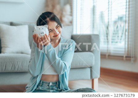 Young Asian woman holding piggy bank and sitting on the floor at home 127673179