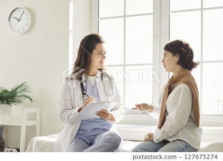Young woman discussing health concerns with doctor while physician taking notes during consultation. Young woman discussing health concerns with doctor while physician taking notes during consultation. 127675274