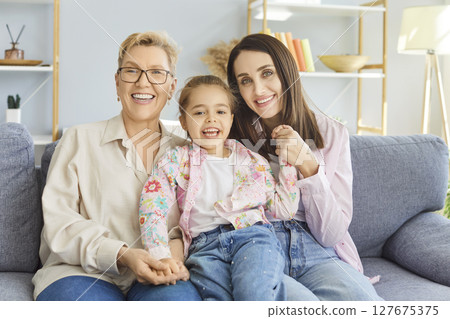 Three generations of smiling women sitting on sofa, hugging and looking at camera during family time 127675375