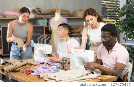 During lesson, woman teacher helps student sew, others work with fabric, blank, dress patterns During lesson, woman teacher helps student sew, others work with fabric, blank, dress patterns 127675435