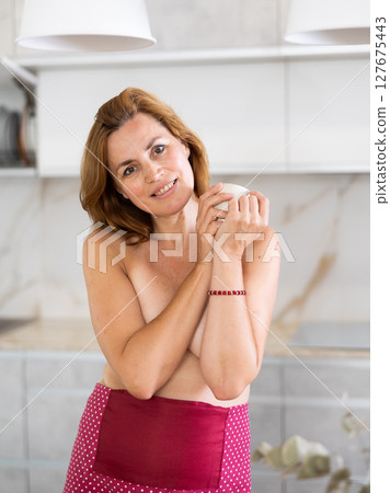 Standing topless woman with apron tied at waist covers bare chest with crossed arms 127675443