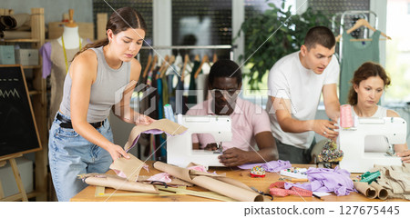 During lesson, guy teacher helps student sew, others work with fabric, blank, dress patterns 127675445