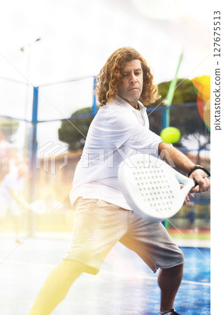 Focused middle-aged Latin man playing padel on court. View through tennis net Focused middle-aged Latin man playing padel on court. View through tennis net 127675513