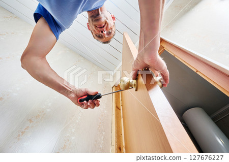 Securing door knobs by tightening screws with screwdriver to fix handles firmly in place inside large predrilled hole in wooden door completing installation by installer. 127676227