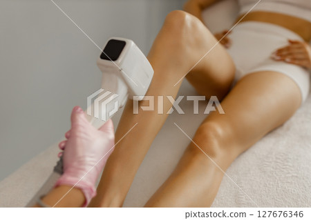 A female cosmetologist does laser hair removal on the slender legs of a beautiful young woman lying on a medical couch in a beauty salon. Cosmetology, hair removal and spa concept. Body care. Close-up 127676346
