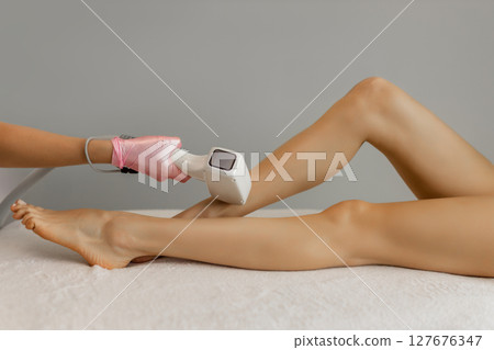 A female cosmetologist does laser hair removal on the slender legs of a beautiful young woman lying on a medical couch in a beauty salon. Cosmetology, hair removal and spa concept. Body care. Close-up 127676347