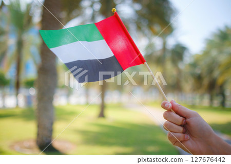 Close up of female hand holding tiny flag of UAE against Abu Dhabi park from Corniche. Promenade in Abu Dhabi. United Arab Emirates, Middle East. Photo of UAE national day celebration spirit. Close up of female hand holding tiny flag of UAE against Abu Dhabi park from Corniche. Promenade in Abu Dhabi. United Arab Emirates, Middle East. Photo of UAE national day celebration spirit. 127676442