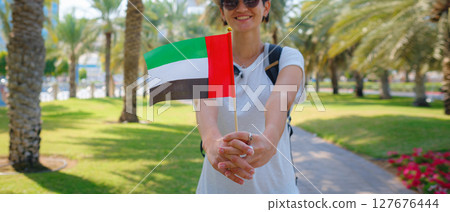 woman holding tiny flag of UAE against Abu Dhabi skyline, Promenade in Abu Dhabi. United Arab Emirates, Middle East. Immigration, student education and Arab emirates citizenship concept 127676444