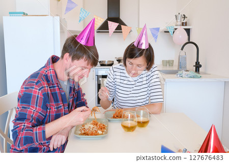 Young couple celebrates cozy birthday at home, enjoying cake and laughter in warm setting. Family are eating pasta and drinking wine to celebrate, enjoy meal and cheers 127676483