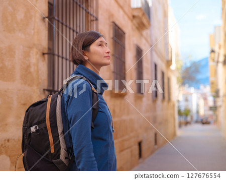 Young woman strolling through charming narrow streets of Denia, Alicante province, Spain, surrounded by historic Mediterranean architecture , enjoying peaceful atmosphere and cultural ambiance. 127676554