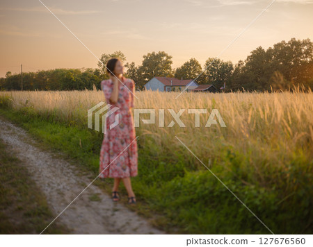 Young woman walking through picturesque European field in late summer. Golden sunlight, lush greenery, and serene rural atmosphere create peaceful countryside scene. Young woman walking through picturesque European field in late summer. Golden sunlight, lush greenery, and serene rural atmosphere create peaceful countryside scene. 127676560