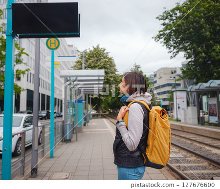 Tourist woman stands at tram stop in Frankfurt, waiting for public transportation amidst urban setting, surrounded by city life and modern buildings, experiencing everyday rhythm of city. 127676600