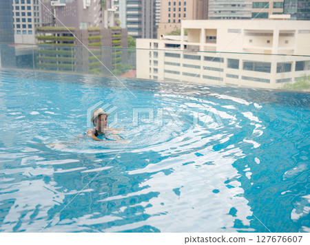 Young asian woman relaxing by pool at Kuala Lumpur hotel with view of surrounding skyscrapers, enjoying leisure time in vibrant urban setting. 127676607