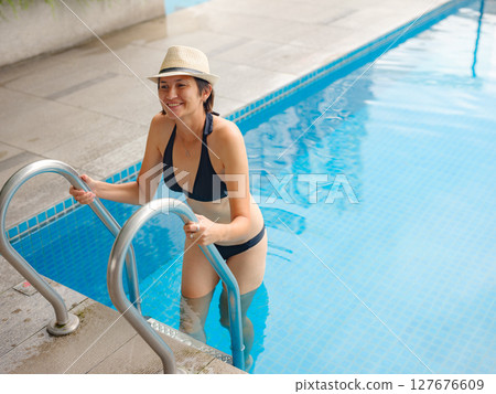 Young asian woman relaxing by pool at Kuala Lumpur hotel with view of surrounding skyscrapers, enjoying leisure time in vibrant urban setting. 127676609