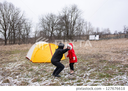 Joyful moment in winter wilderness as child in red coat plays with adult near cozy yellow tent under cloudy skies 127676618