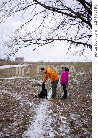 Couple enjoys a winter stroll with their dog on a snowy path under a bare tree in a serene landscape during a chilly afternoon 127676690